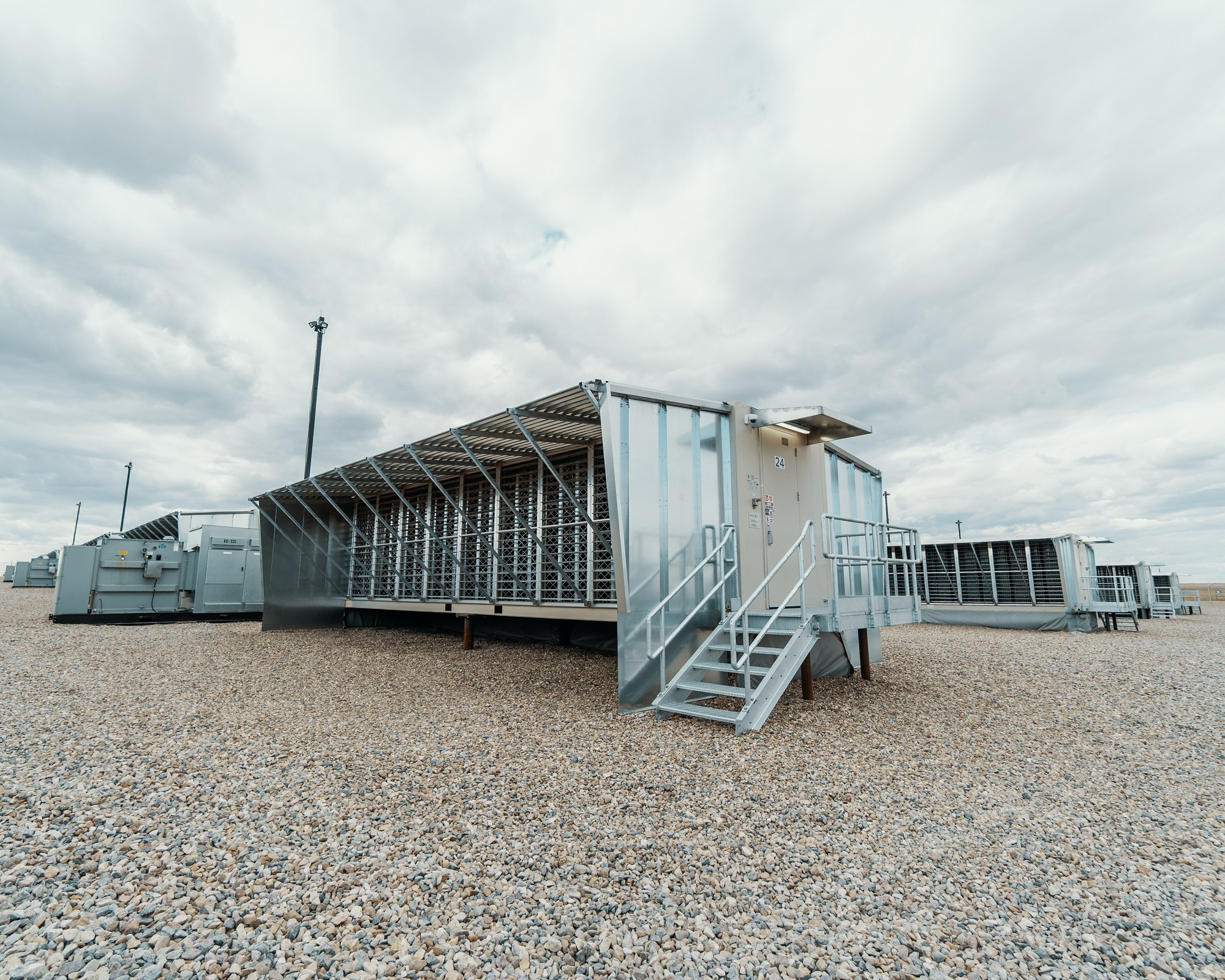 A Hut 8 mining unit at ground level under an overcast prairie sky — an open-sided steel container of fan louvers and racks, with rows of others behind it.