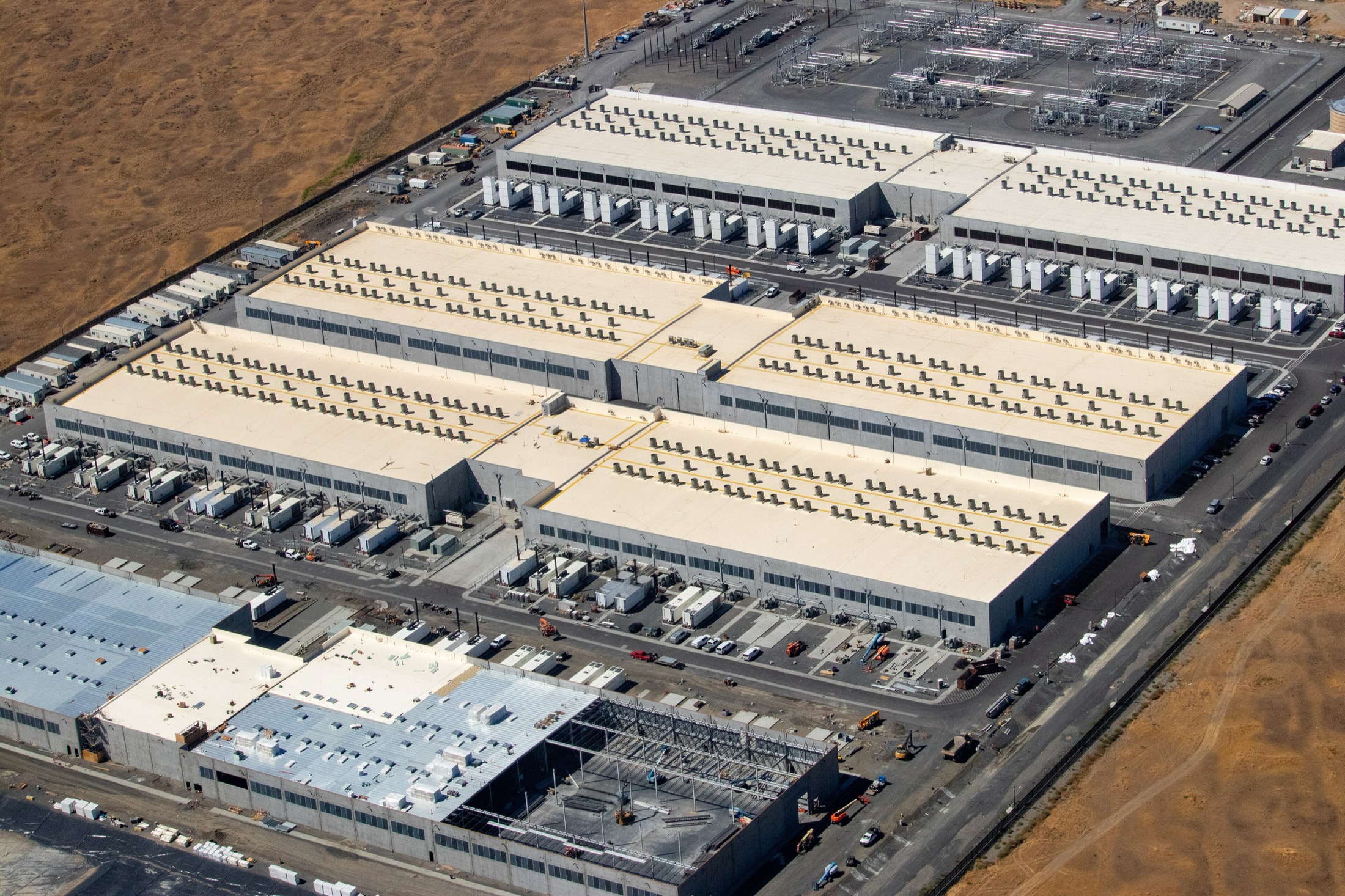 Aerial view of a hyperscale data-center campus in Oregon: three finished white halls with rooftop cooling, a fourth in exposed steel, and a substation switchyard at the edge.