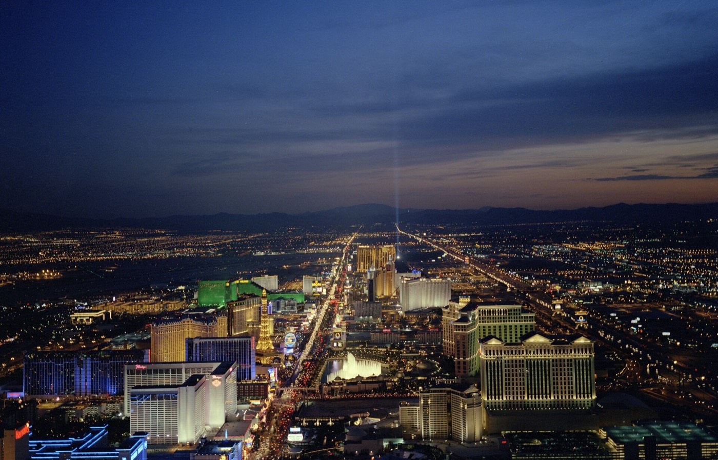 Elevated dusk view down the length of the Las Vegas Strip corridor, city lights stretching to the horizon and a vertical searchlight beam mid-frame.