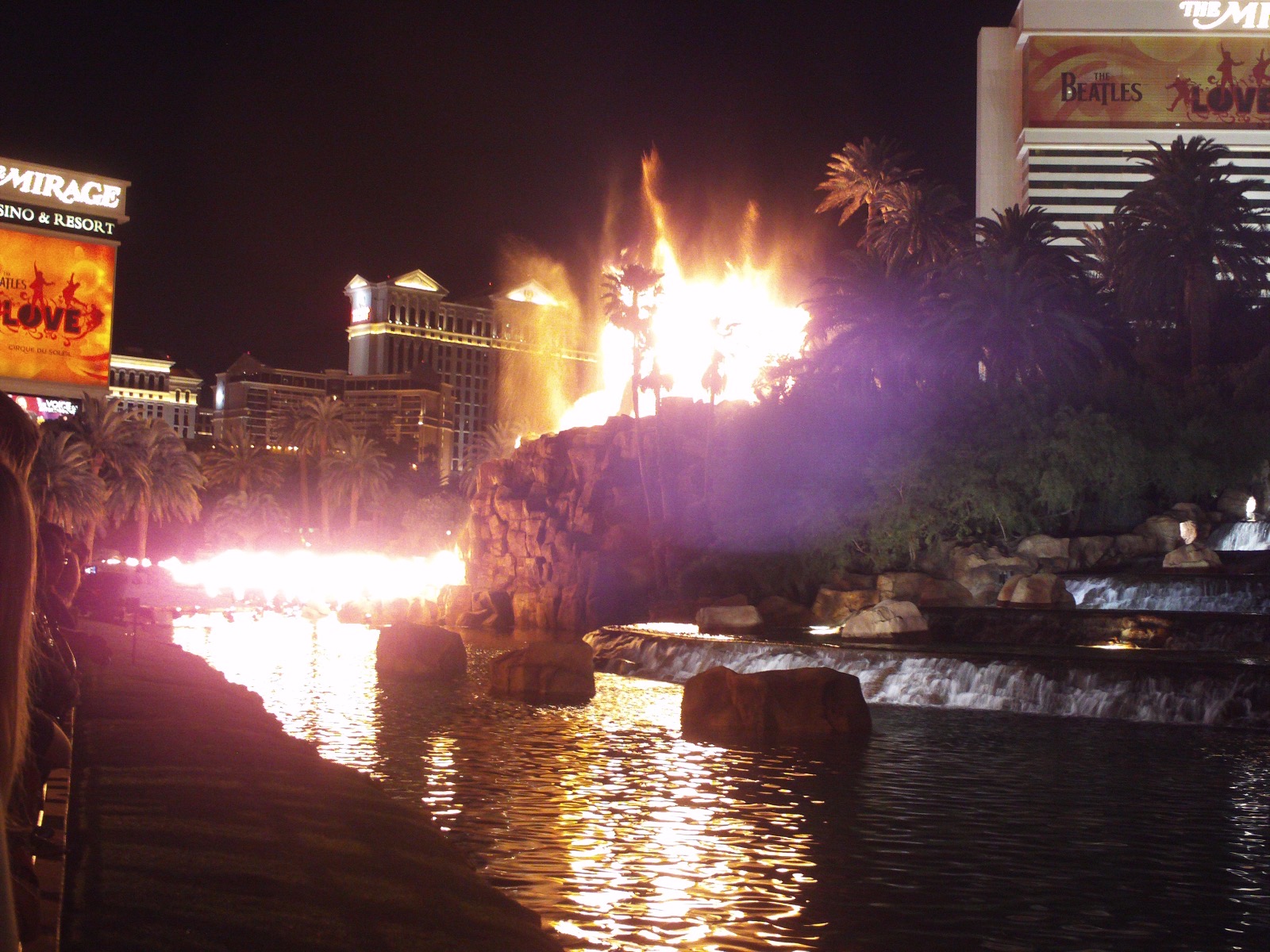 The Mirage’s volcano attraction erupting at night, flames and mist rising over the rock formation, with the resort’s sign at left.