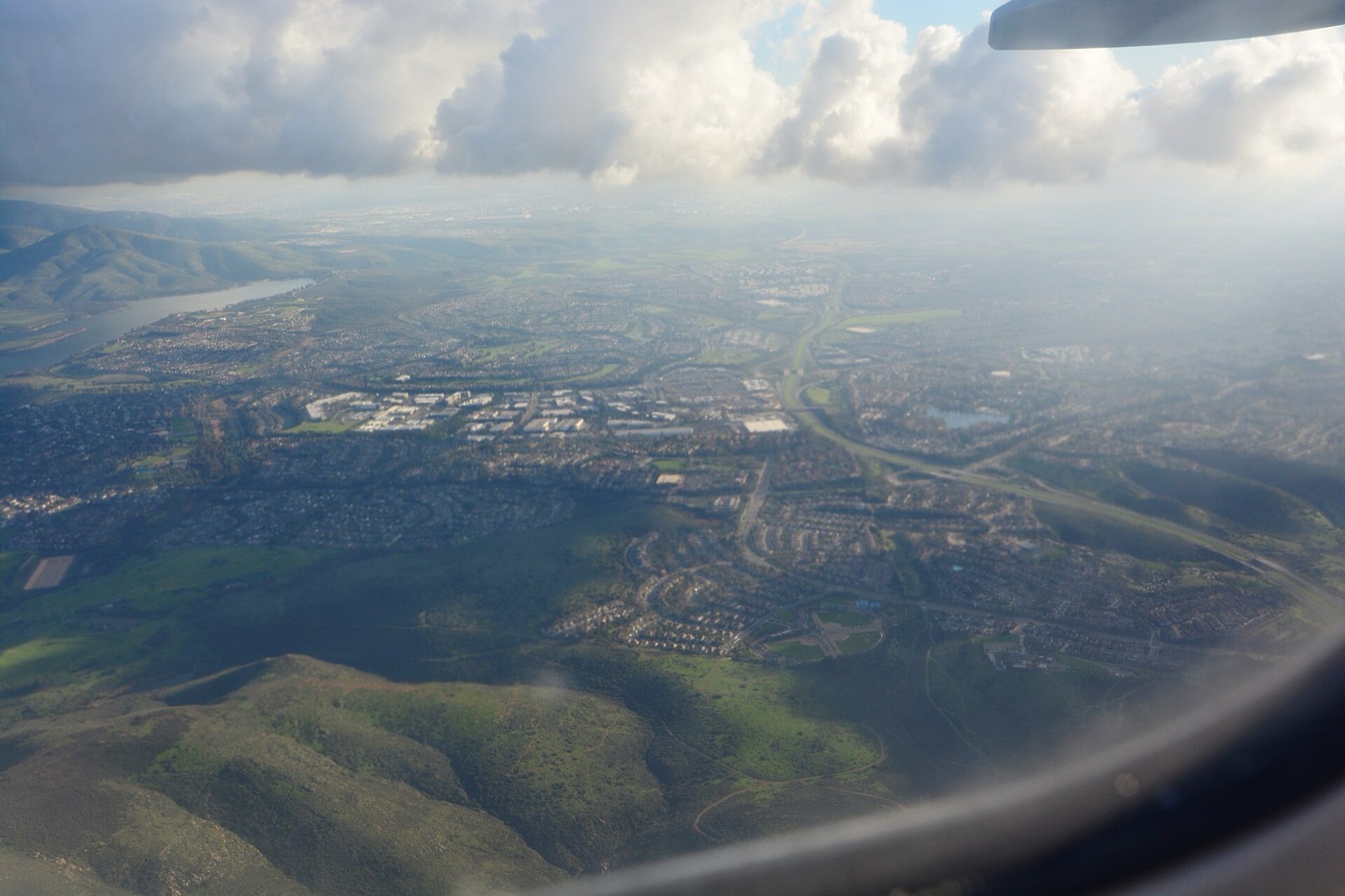 Aerial view looking south over Chula Vista from a small plane: Lower Otay Lake at upper left, dense subdivision streets filling the middle distance, and undeveloped chaparral hills in the near foreground.