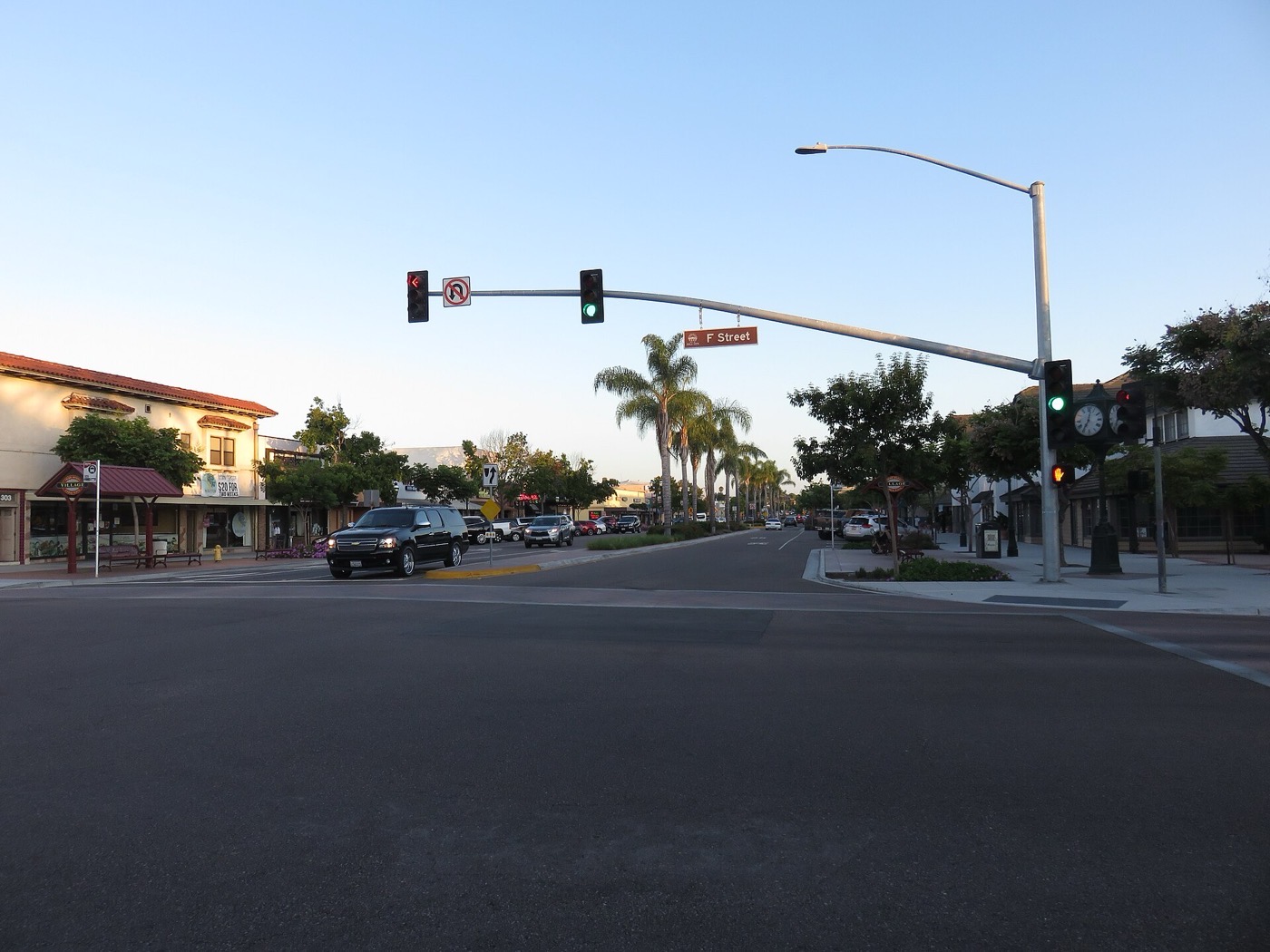A street intersection in downtown Chula Vista, with traffic signals, palm trees and low-rise storefronts at dusk.