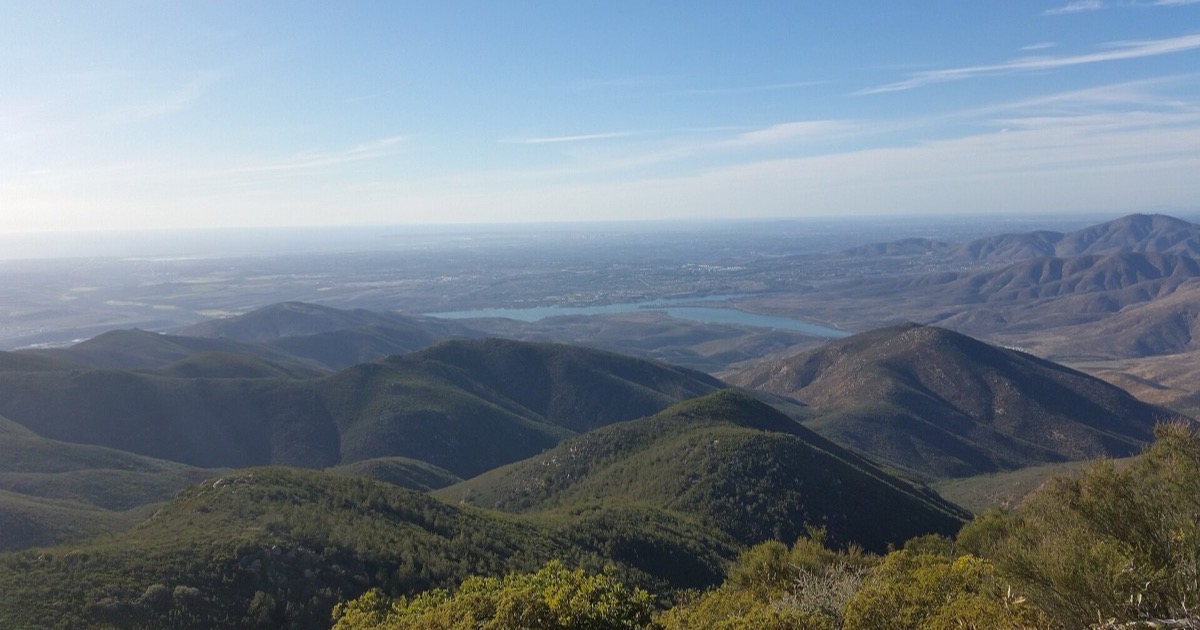 Undeveloped chaparral-covered hills in south San Diego County, with a reservoir visible in the valley below and hazy urban development on the far horizon.