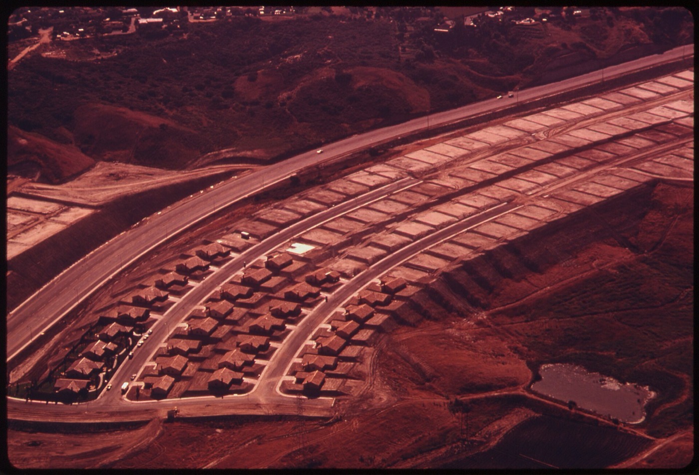 Aerial view of a 1970s tract-housing development under construction, with finished houses on curving streets and raw graded lots still open on the hillside beyond.
