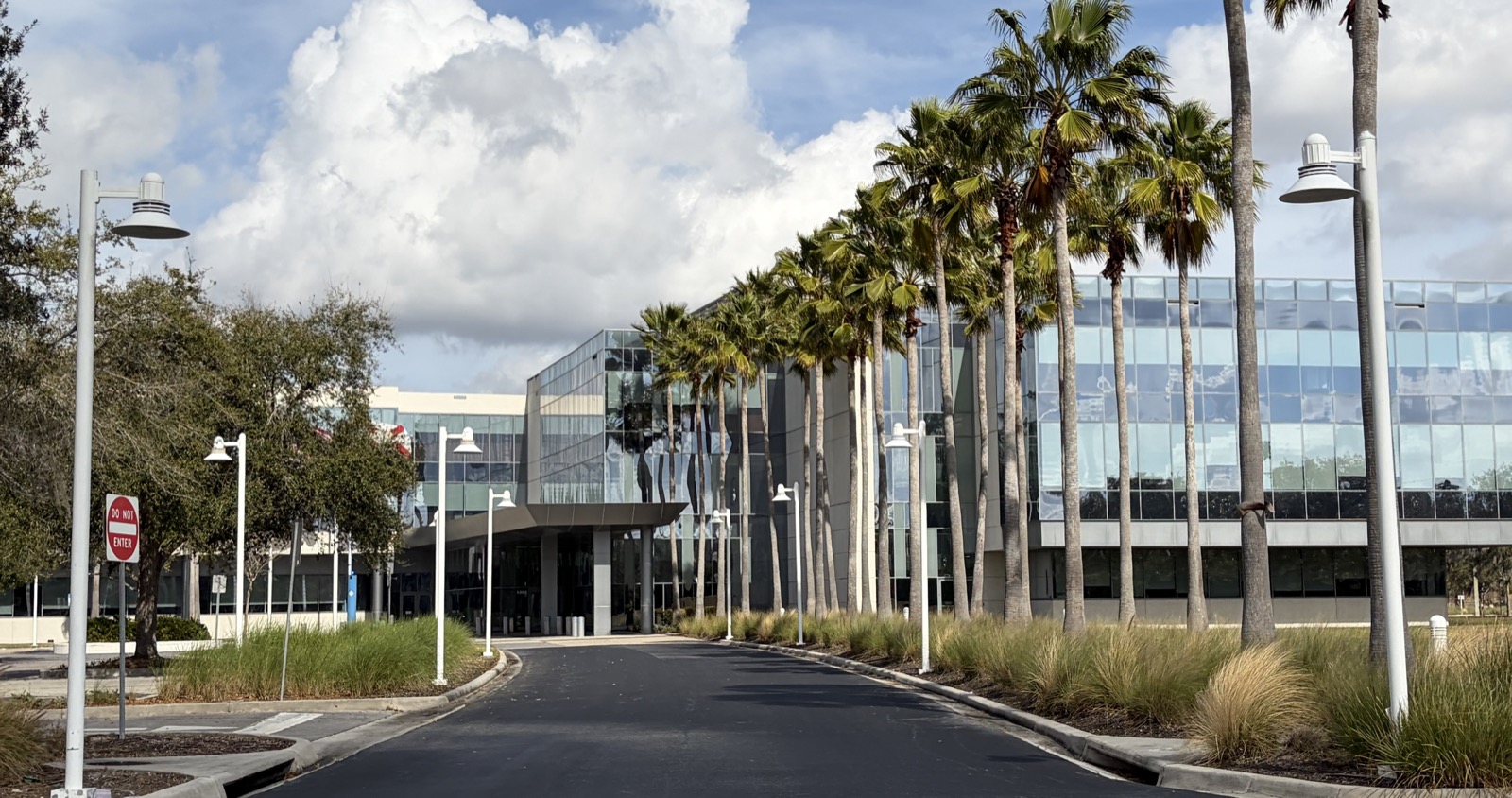The glass office building of Darden Restaurants’ corporate headquarters campus near Orlando, Florida, viewed along a palm-lined driveway under a partly cloudy sky.