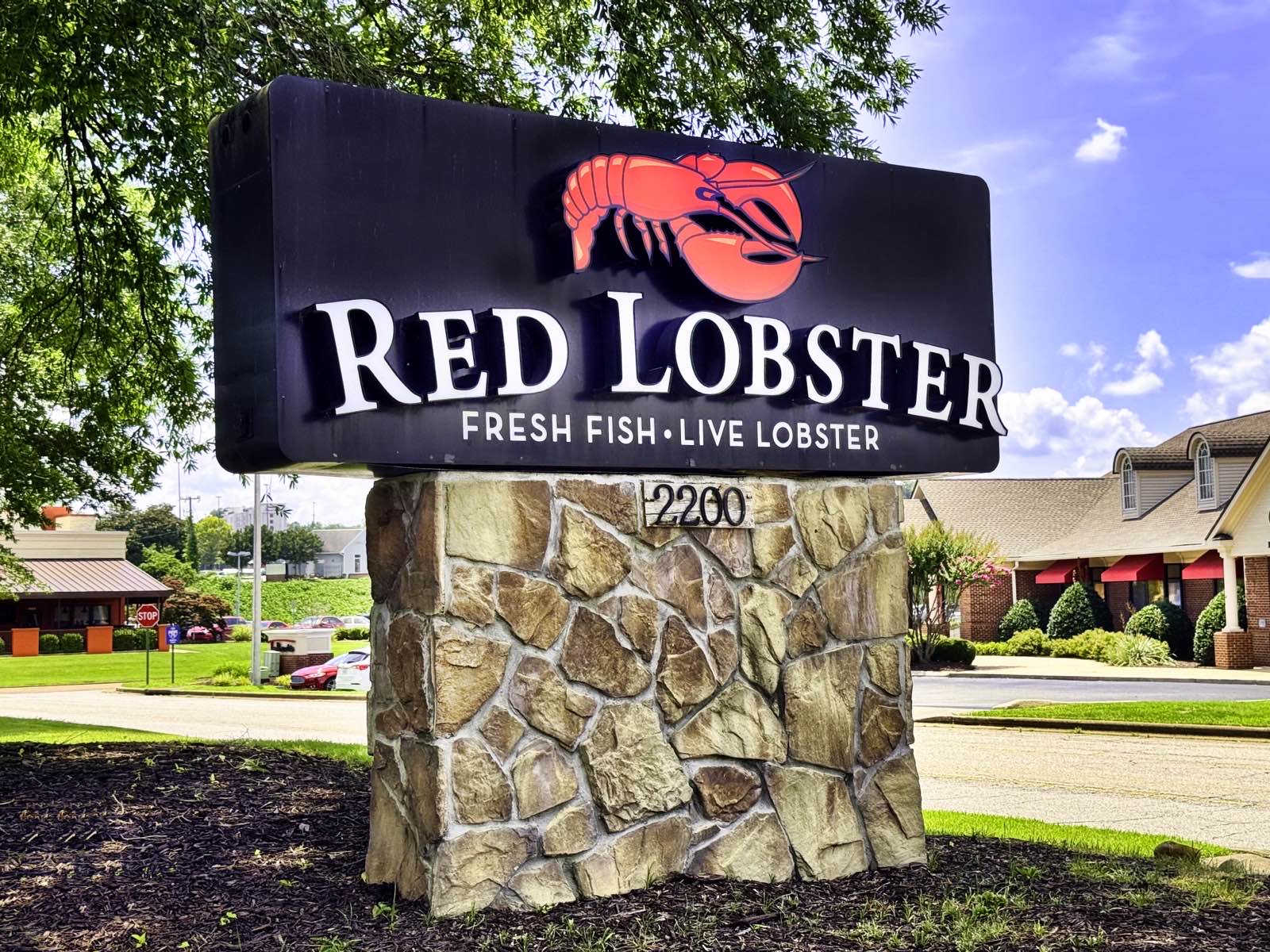 A close-up of a Red Lobster roadside sign on a stone base, showing the lobster mascot, the RED LOBSTER wordmark and the line FRESH FISH · LIVE LOBSTER against a blue sky.