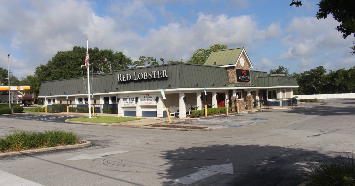 A freestanding Red Lobster restaurant with a green metal roof and a Red Lobster sign on the front gable, photographed from its parking lot under a partly cloudy blue sky.