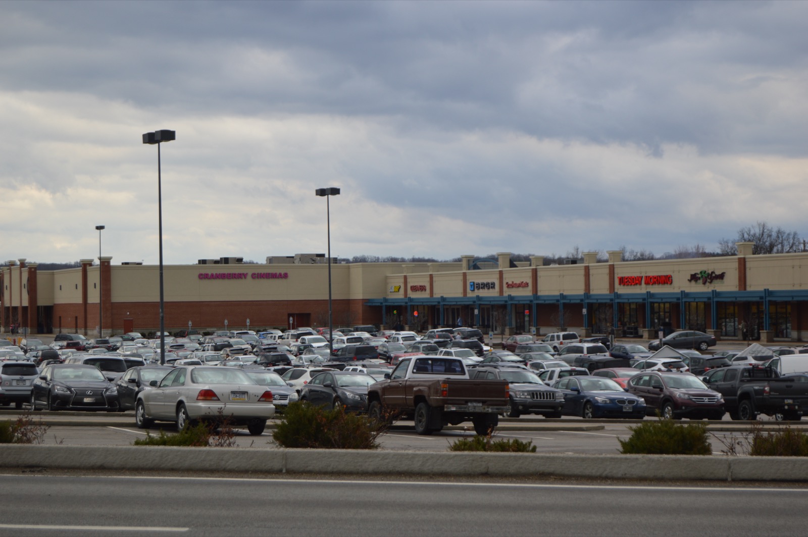 A wide view of a suburban shopping-mall parking lot filled with cars, a strip of big-box storefronts and a cinema sign behind it under an overcast sky.