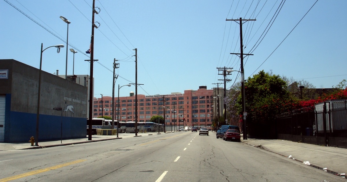 The intersection of Seventh and Alameda in downtown Los Angeles before redevelopment — the pink American Apparel factory building rising behind the Greyhound maintenance yard, power lines crossing the street.