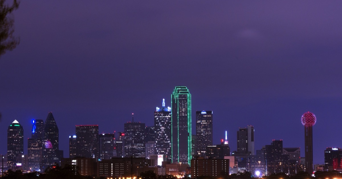 The downtown Dallas skyline at dusk under a purple sky — Bank of America Plaza outlined in green light, Reunion Tower glowing at the right.