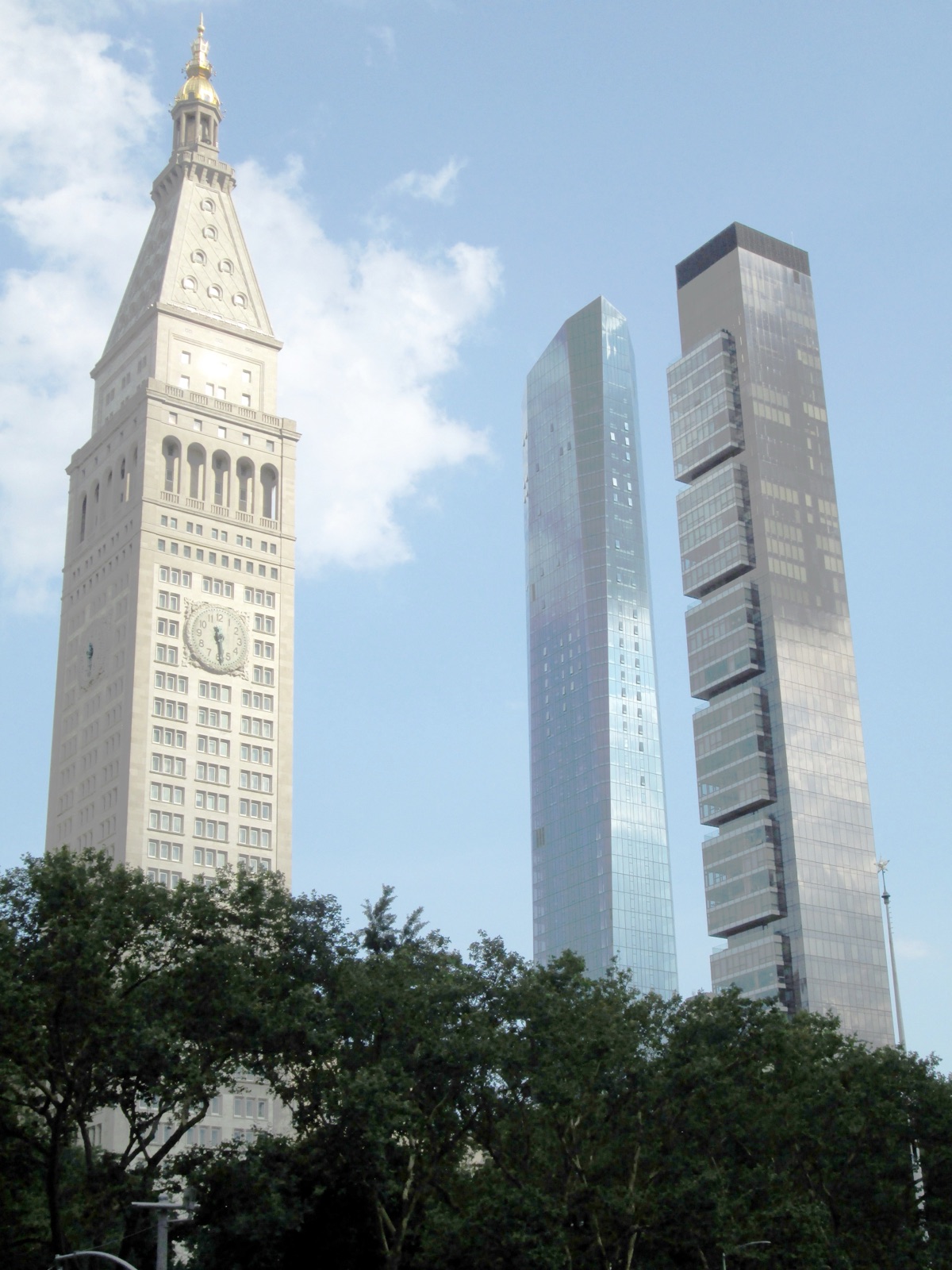 The ornate cream-colored Metropolitan Life Insurance Company clock tower stands beside the glass twin towers of One Madison Avenue on the former MetLife North Building site, seen above trees against a blue sky.