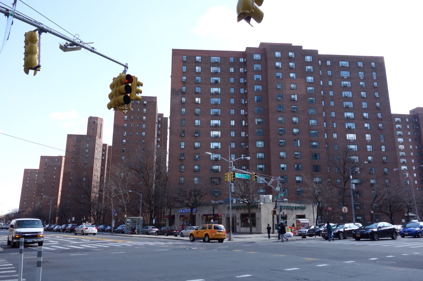 Brick high-rise towers of Peter Cooper Village rise behind a Manhattan street corner with ground-floor shops, a yellow taxi, traffic signals, and parked cars, photographed in winter with bare trees.