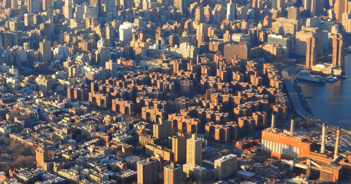 Aerial view at golden hour of dozens of near-identical reddish-brown brick apartment towers arranged in a grid pattern within Stuyvesant Town–Peter Cooper Village, with the East River, FDR Drive, and a power plant to the right and the Midtown Manhattan skyline visible in the hazy distance beyond.