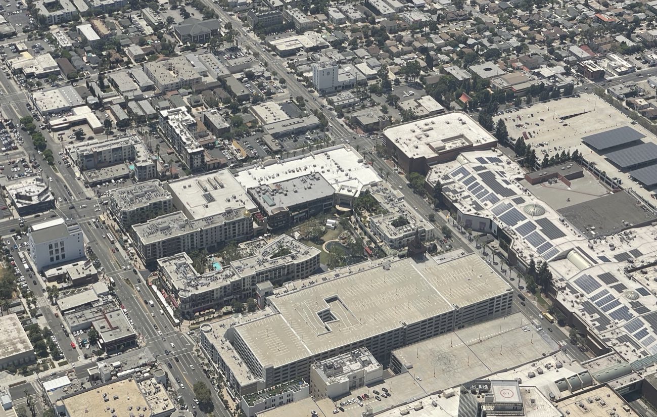 An aerial photograph of downtown Glendale, California, showing the open blocks and central green of the Americana at Brand on the left, apartments stacked above its shops, and the large solar-panelled roof of the enclosed Glendale Galleria directly across the street on the right.