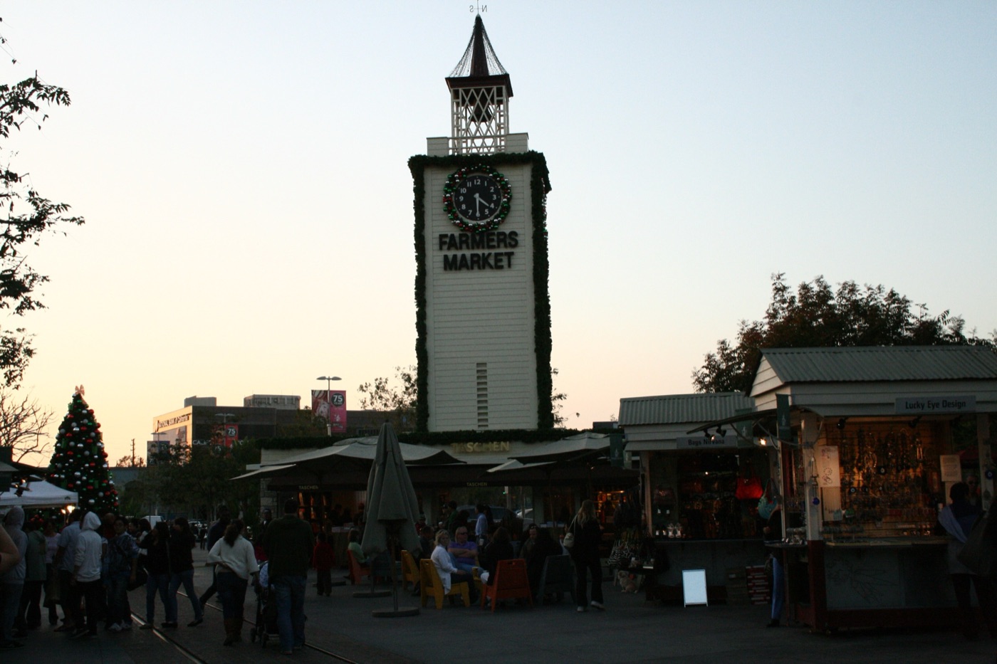 The Original Farmers Market's white clock tower at dusk in November 2009, hung with holiday garlands and banners reading 75 Years, with market stalls below and the trolley rails running through the foreground.