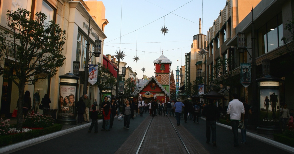First Street at The Grove in Los Angeles at dusk in November 2009, with the trolley's steel rails running down the centre of the paved street, shoppers walking on either side, the Barnes & Noble and The Grove blade signs above, and a holiday village at the far end.