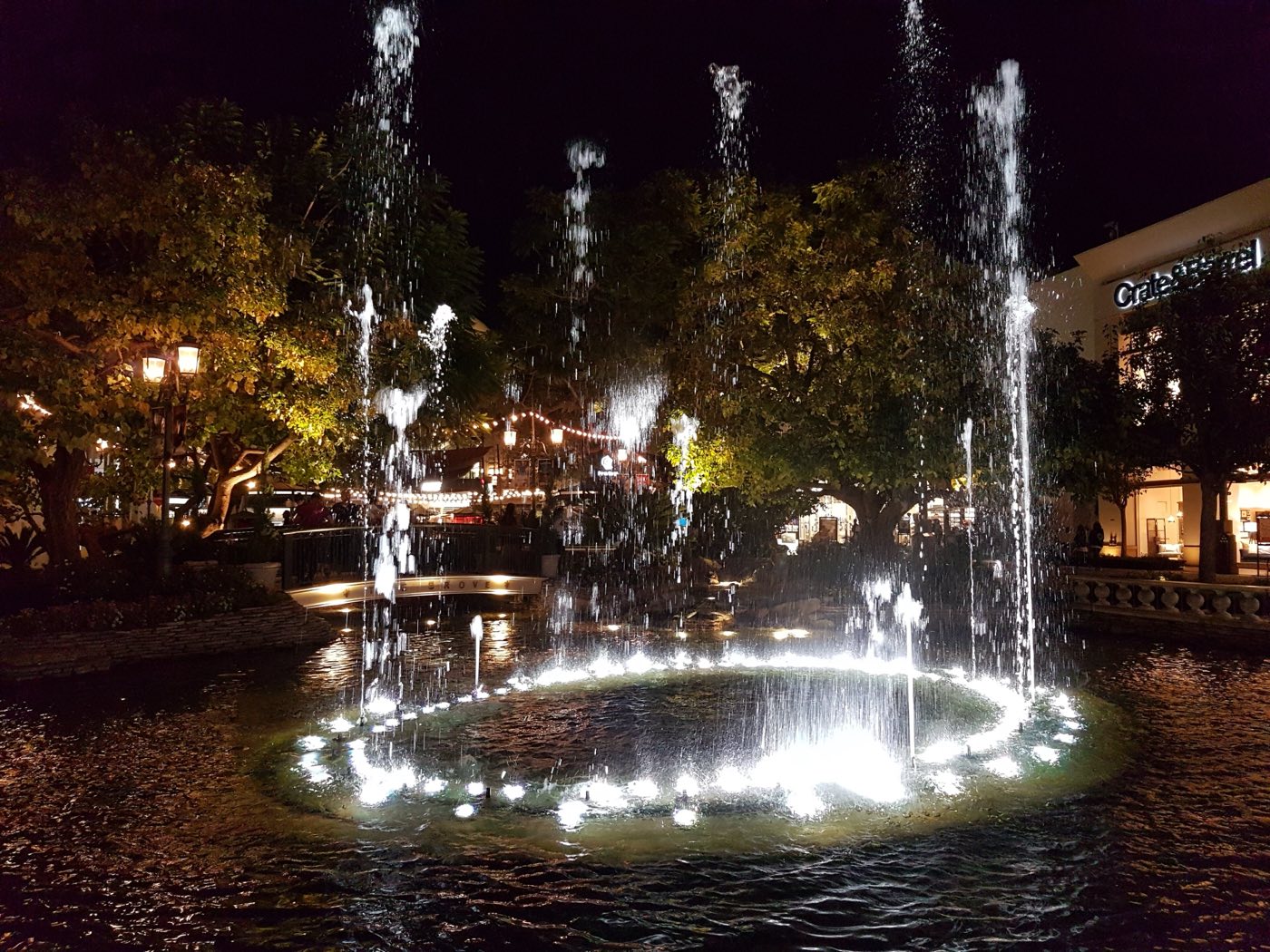 The Grove's lit dancing fountain at night in September 2016, jets rising from a floodlit ring in the pool, with a low bridge lettered THE GROVE behind it, string lights in the trees and a Crate & Barrel storefront to the right.