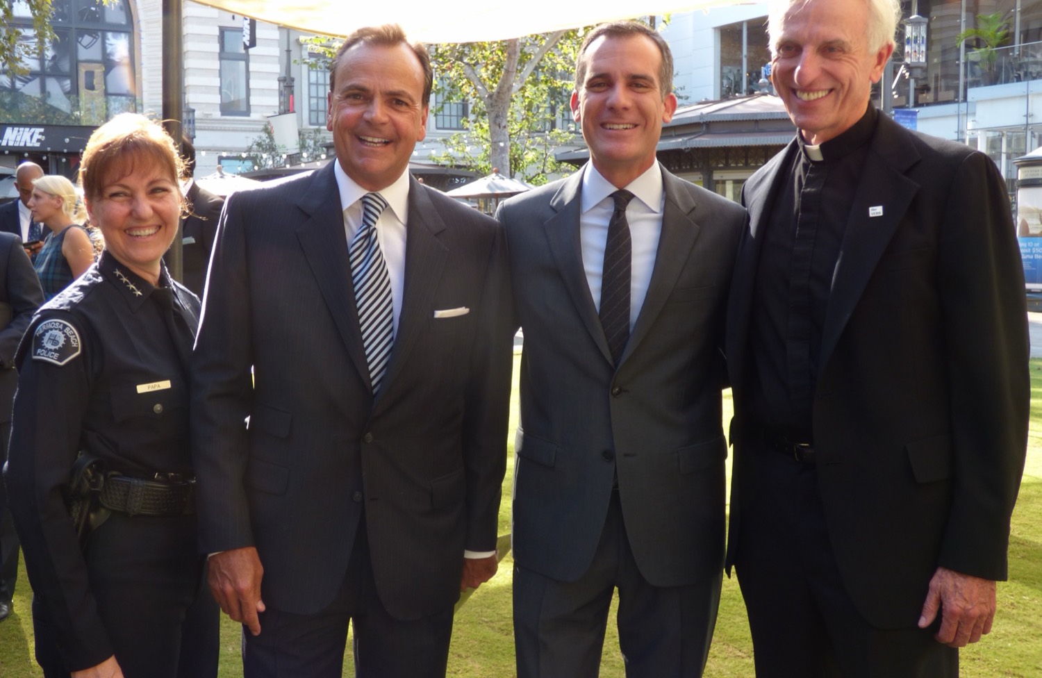 Four people standing on a lawn under a canopy in September 2015 — a uniformed Los Angeles police officer, the developer Rick Caruso in a dark suit and striped tie, a man in a grey suit, and a priest in a clerical collar.