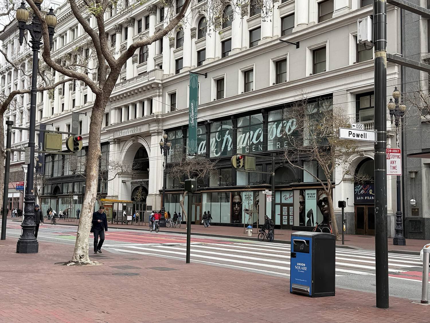 The older Market Street frontage of San Francisco Centre at Powell Street in February 2026 — a pale classical facade with columns and round-arched openings, San Francisco Centre in script across the glazing, and a shuttered Ray-Ban shopfront papered with advertising below.