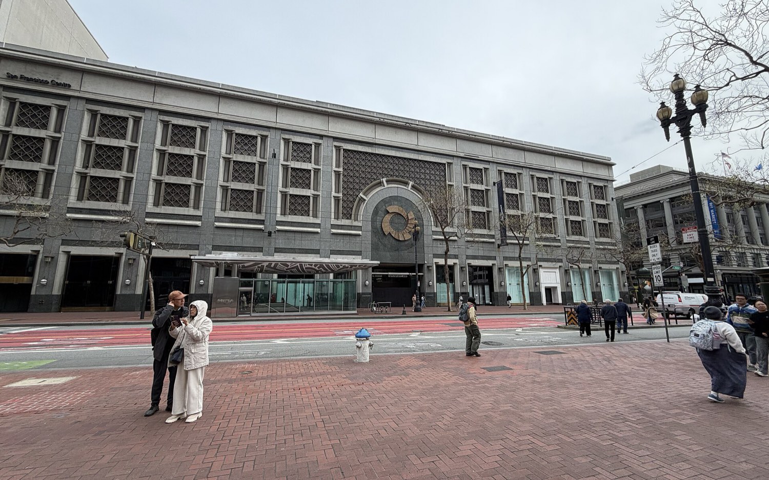 The Market Street front of San Francisco Centre on 14 February 2026 under a grey sky — the same granite facade with all retail signage stripped, the entrance fenced off, and a near-empty brick plaza in front.