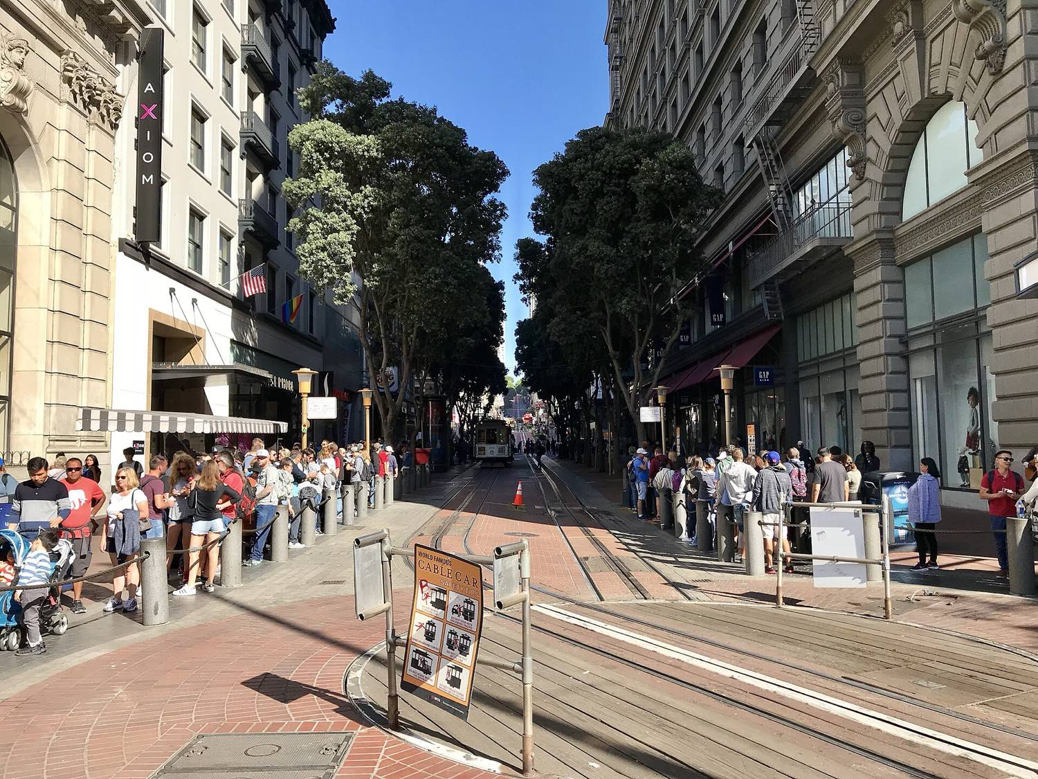 The cable car turntable at Powell and Market in October 2017 — two lines of waiting passengers roped off on either side of the wooden turntable, a cable car standing further up Powell Street between plane trees, under a clear blue sky.