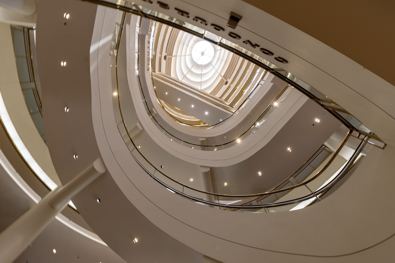 Looking up through the west atrium of San Francisco Centre in July 2023 — nested rings of curved balconies and spiral escalators rising to a lit oval skylight, with reversed NORDSTROM lettering across the top of the frame.