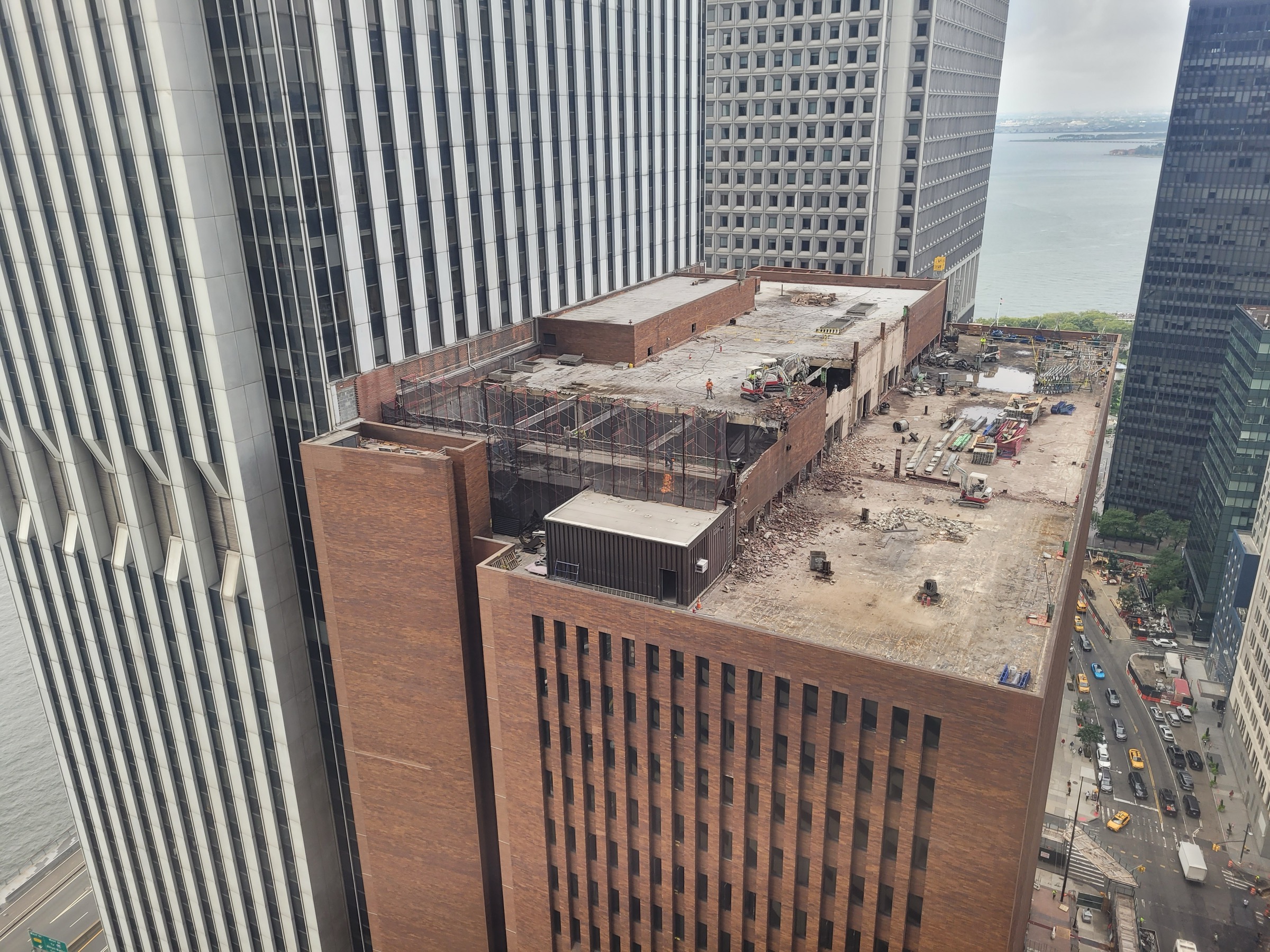 The roof of 25 Water Street in August 2023, seen from above: excavators tearing open the brick crown of the 1969 tower, the harbor beyond, demolition debris across the deck.