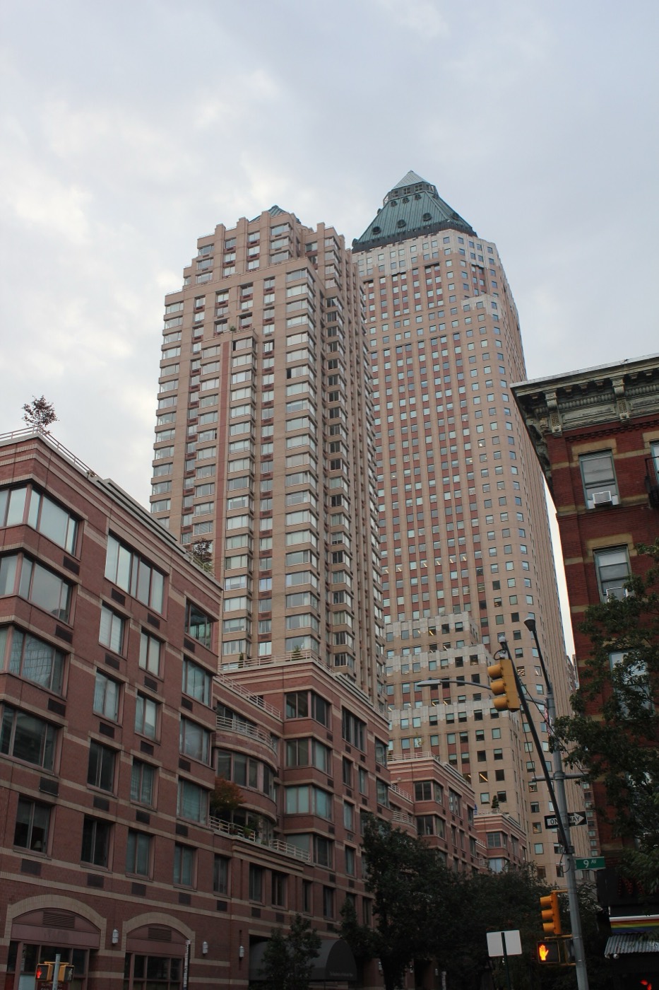The Worldwide Plaza complex in Manhattan seen from street level on Ninth Avenue — brick residential buildings in the foreground and a tall stone office tower with a copper pyramid crown rising behind them, under an overcast sky.
