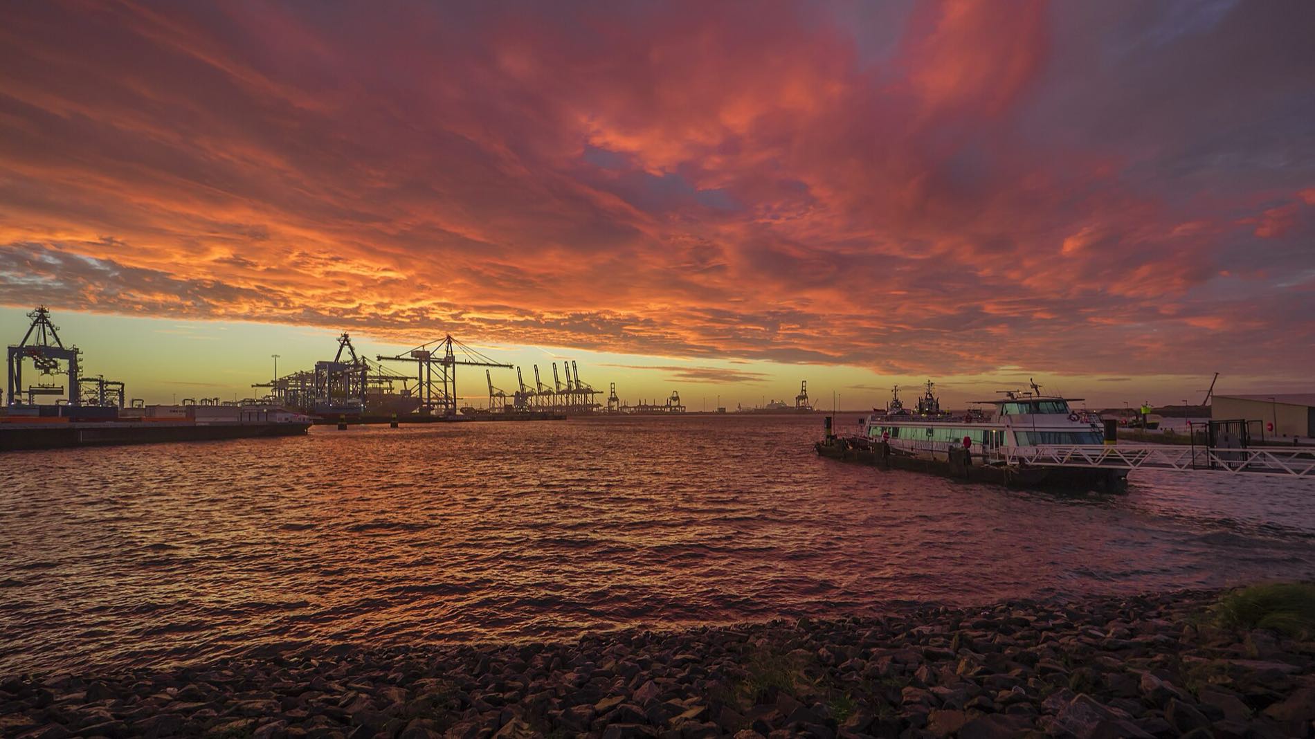 Container gantry cranes standing in silhouette along the far quay of a deep-water harbour at sunset, an orange and violet sky filling the frame above, a moored ship and a small ferry on the water, and stacked containers at the left edge.