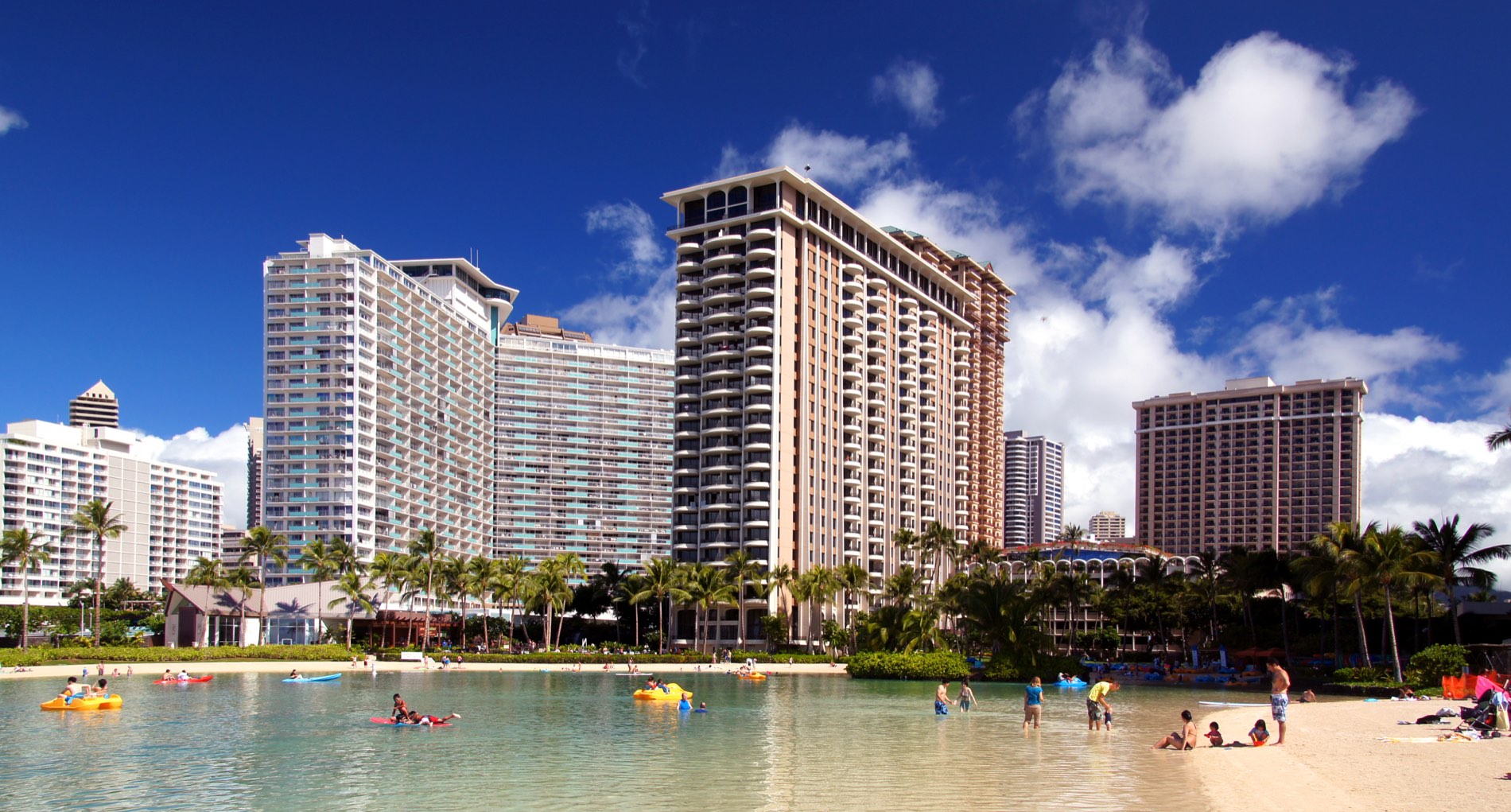 The Hilton Hawaiian Village on Waikiki seen across the Duke Kahanamoku lagoon: a row of tall balconied resort towers behind a line of palms, pedal boats and swimmers in green water, and a white sand beach under a deep blue sky.