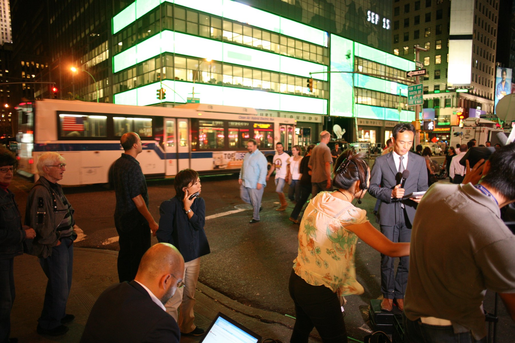 Lehman Brothers' Seventh Avenue headquarters at night, its glass facade lit in bands of green and white with a screen reading SEP 15, a city bus crossing in front of it and television crews filming to camera on the pavement below.