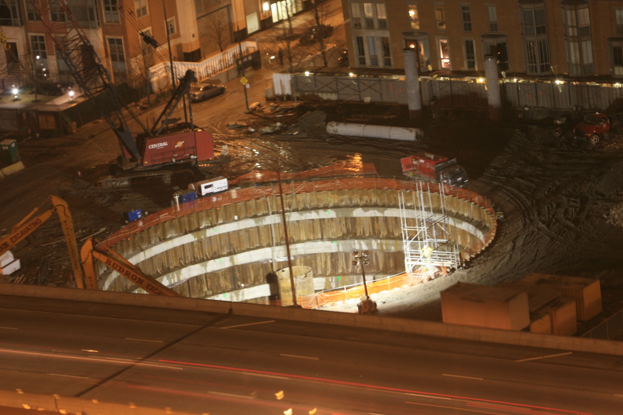 The Chicago Spire excavation at night in January 2008, the terraced circular walls lit by work lights, excavators at the rim and traffic streaking past on Lake Shore Drive.