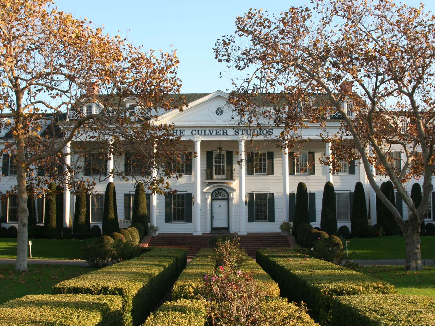 The white colonnaded mansion of The Culver Studios in Culver City in late afternoon light, its portico lettered THE CULVER STUDIOS, with clipped hedges and rose beds on the lawn in front and bare plane trees on either side.