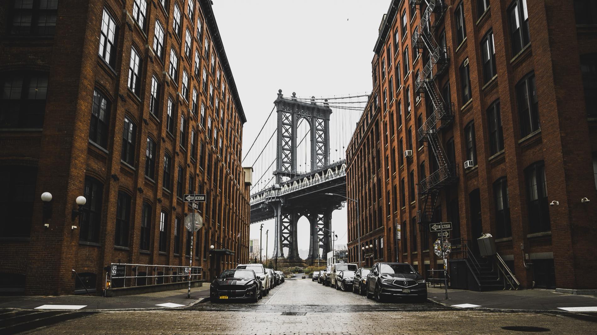 Looking down Washington Street in DUMBO on a wet grey morning — tall red-brick warehouse walls with fire escapes closing both sides of the frame, and a stone-and-steel tower of the Manhattan Bridge standing at the end of the street.