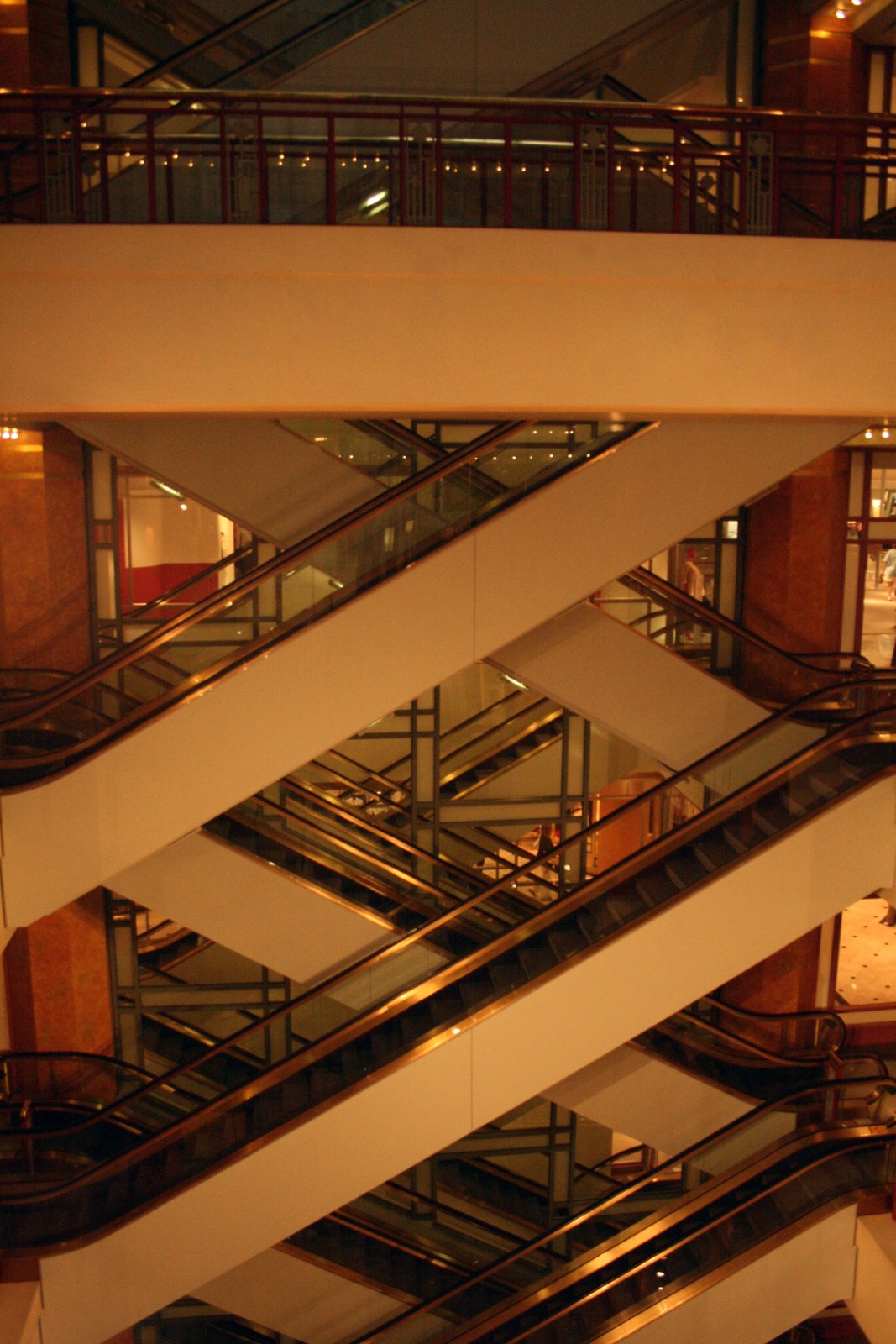 A vertical view down through a mall atrium showing criss-crossing escalators on multiple levels under warm lighting.
