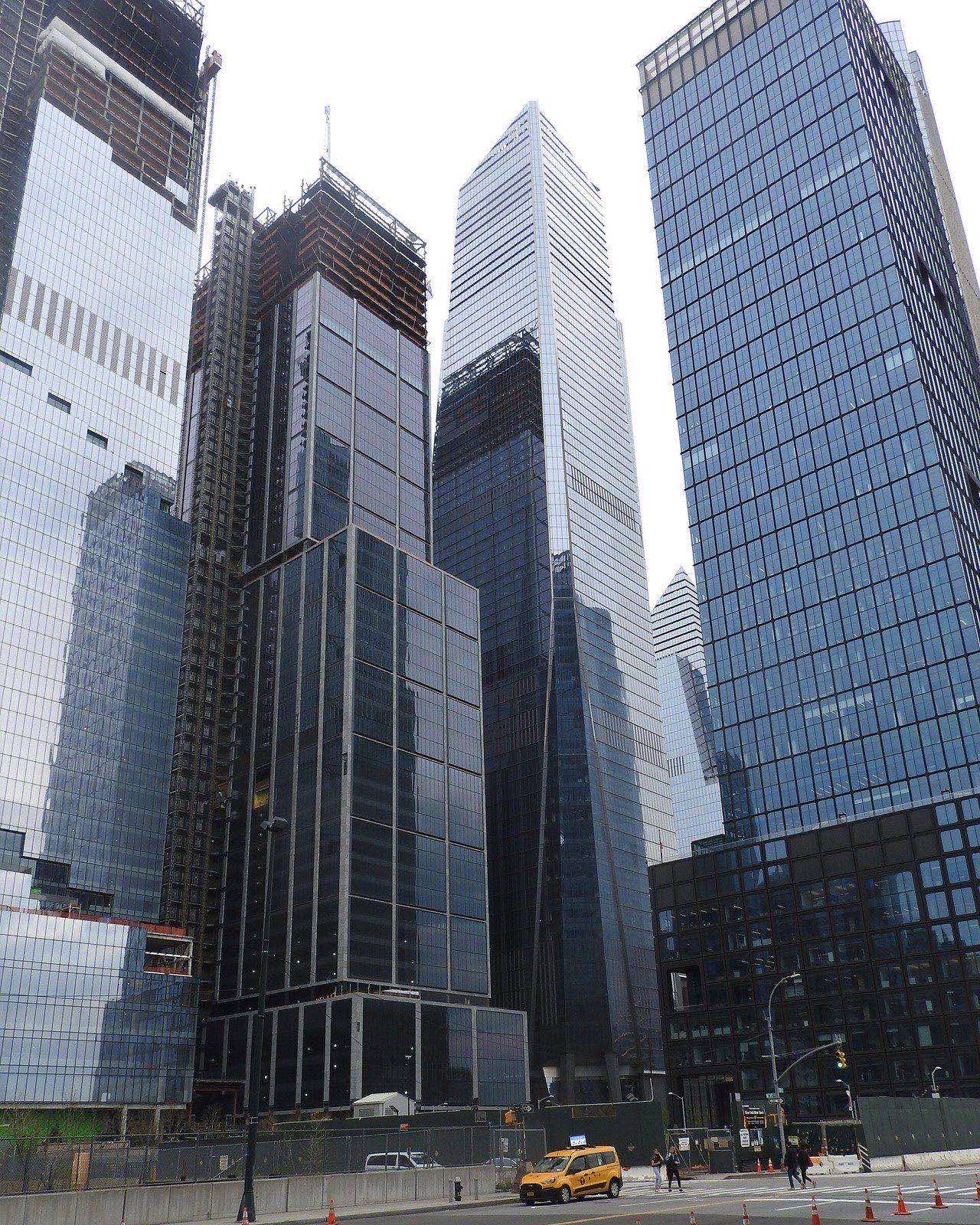 Upward view of glass Hudson Yards towers, one still wrapped in construction scaffolding, forming a narrow canyon above a street with a yellow taxi.