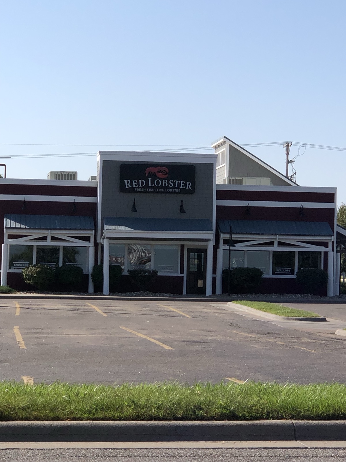 A Red Lobster restaurant photographed from its parking lot in portrait orientation — a low dark-red building with a wall sign reading Red Lobster, Fresh Fish, Live Lobster, under a large expanse of blue sky.