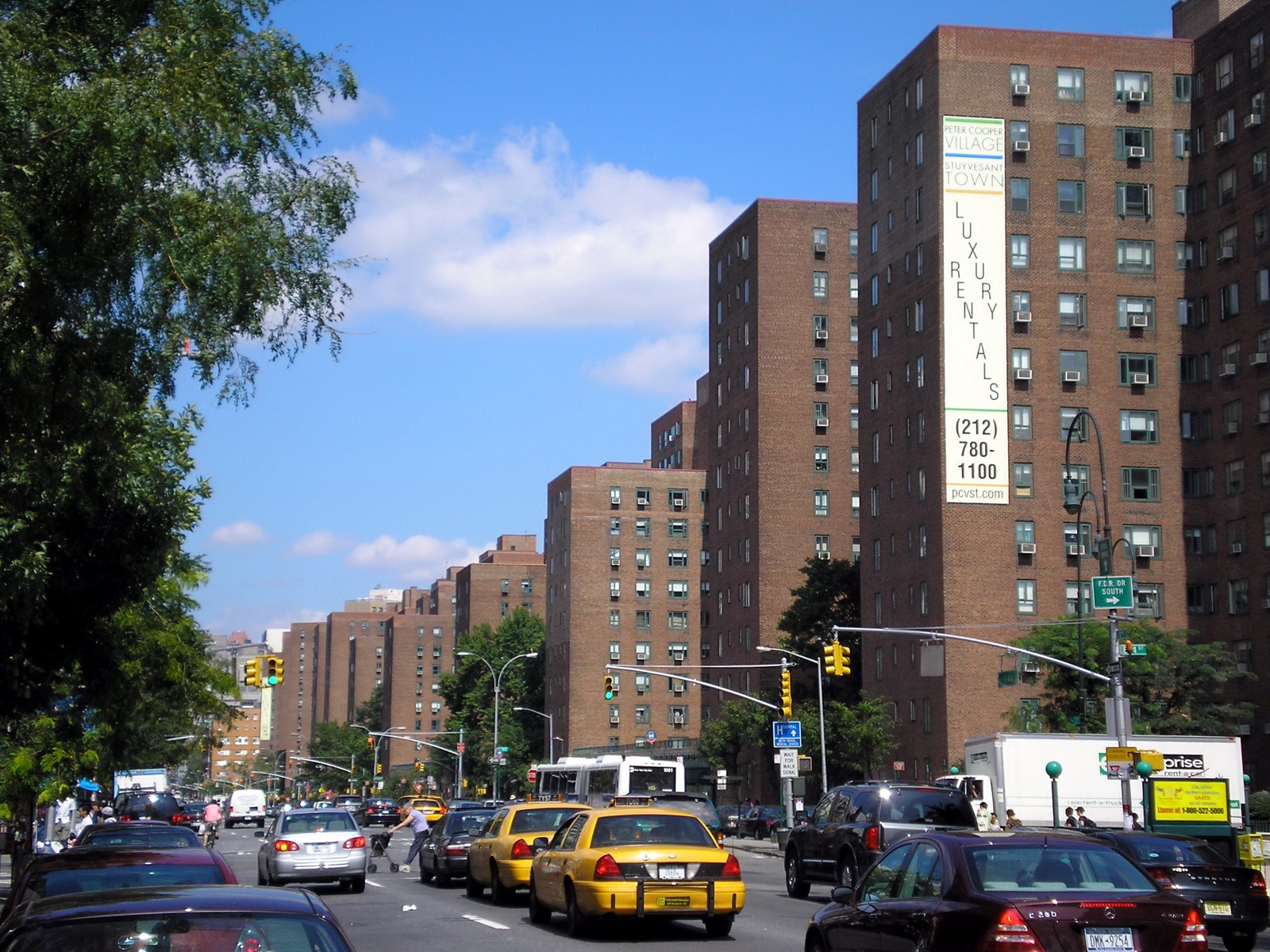 A First Avenue street scene with taxis and traffic; a tall brick apartment tower at right carries a large painted sign reading Peter Cooper Village, Stuyvesant Town, Luxury Rentals, with a phone number and website.