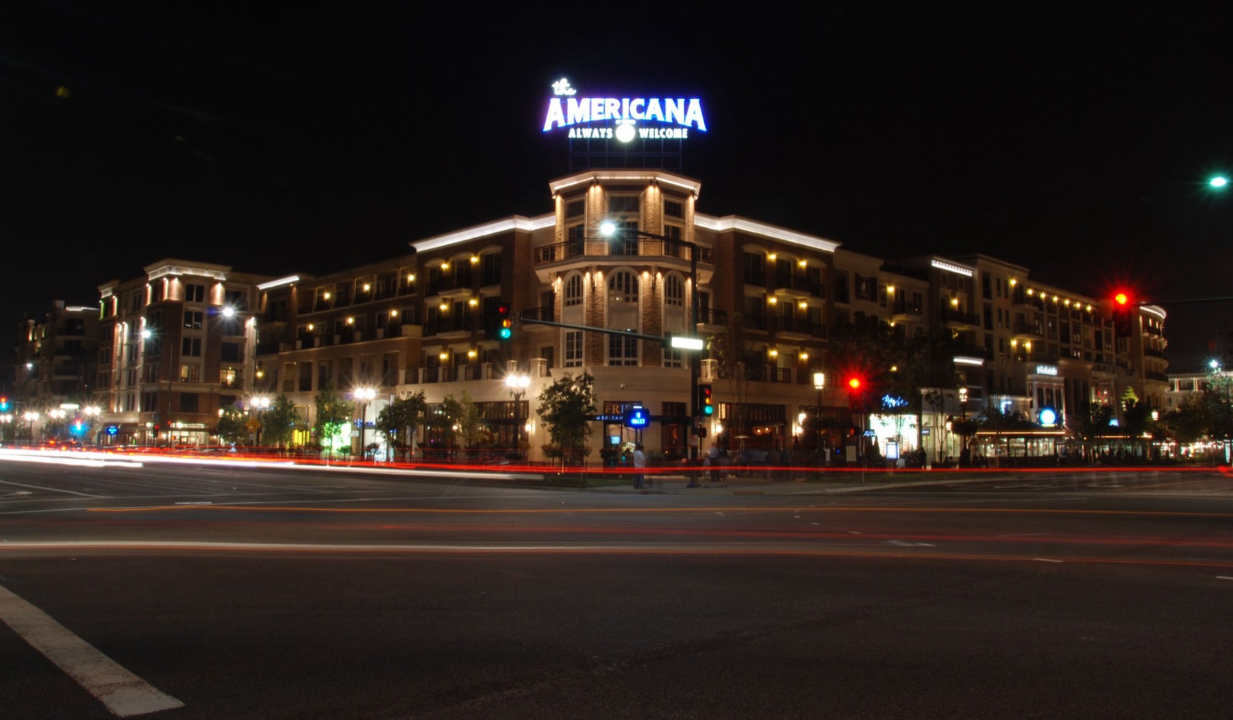 The Americana at Brand in Glendale at night in August 2008, its illuminated rooftop sign above floors of apartments stacked over lit shopfronts on the corner, with red and white car-light trails streaking across the empty intersection in the foreground.