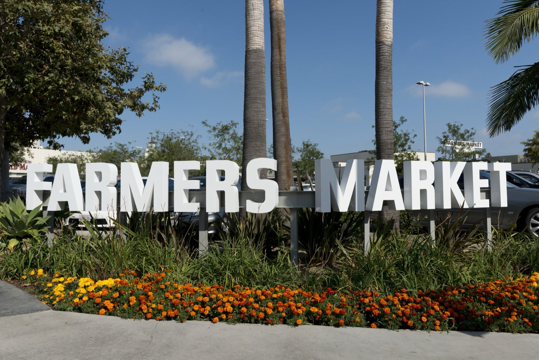 The white block-letter FARMERS MARKET sign standing in a bed of orange and yellow marigolds at Third and Fairfax in Los Angeles, palm trunks and parked cars behind it under a clear blue sky.