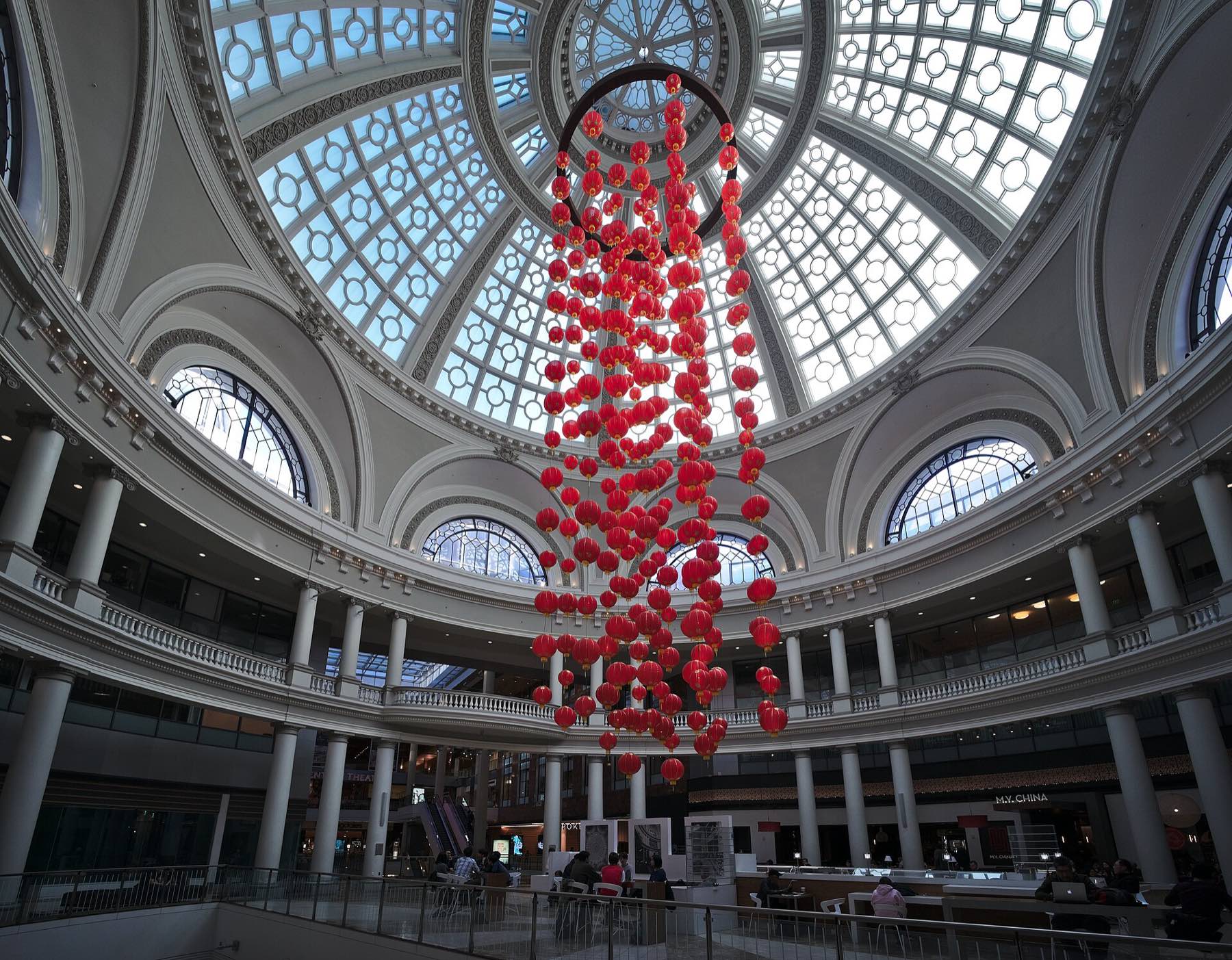 Looking up into the restored Emporium dome inside San Francisco Centre — a coffered glass-and-steel cupola over a ring of columned balconies, with hundreds of red paper lanterns hanging in a single column from its centre down to the food court below.